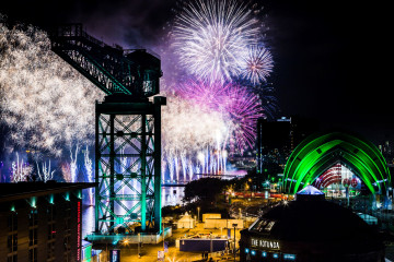 A vibrant night scene in Glasgow with fireworks illuminating the sky in white, purple, and pink over the Clyde Arc bridge and Finnieston Crane. The green-lit SEC Armadillo, Hilton Garden Inn, and The Rotunda add to the festive atmosphere.