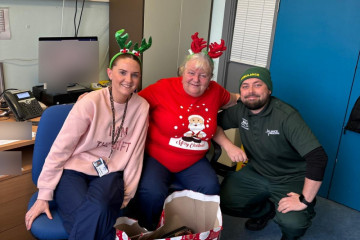 Alliance Ambulance and NHS Scotland team members share a festive moment in an office, wearing Christmas-themed attire and reindeer headbands. Gift bags filled with holiday items surround them, highlighting the joyful spirit and team camaraderie.