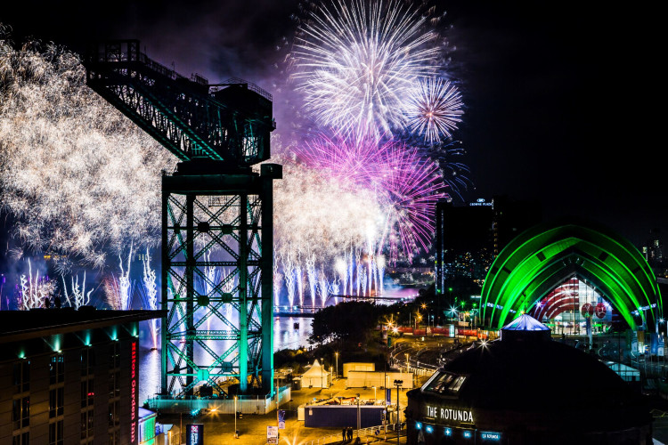 A vibrant night scene in Glasgow with fireworks illuminating the sky in white, purple, and pink over the Clyde Arc bridge and Finnieston Crane. The green-lit SEC Armadillo, Hilton Garden Inn, and The Rotunda add to the festive atmosphere.