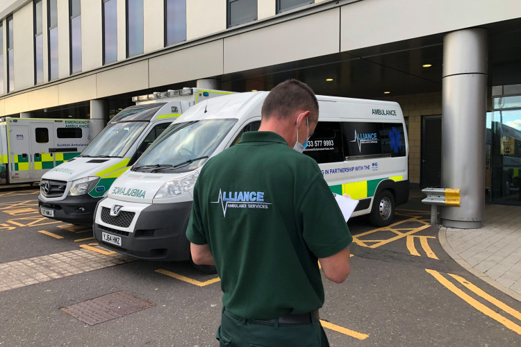 A member of the Alliance Ambulance Services staff, wearing a green uniform, is seen from behind while standing in front of multiple ambulances parked outside a hospital building in Scotland. The ambulances are branded with NHS Scotland and one has an Alliance Ambulance Service logo.