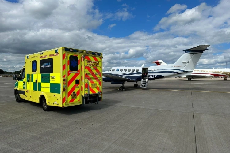 A yellow emergency ambulance from Alliance Ambulance Services is parked on the tarmac near a private air ambulance jet at an airport, under a partly cloudy sky. The vehicles are prepared for medical transportation, with the jet's door open and ready for boarding.
