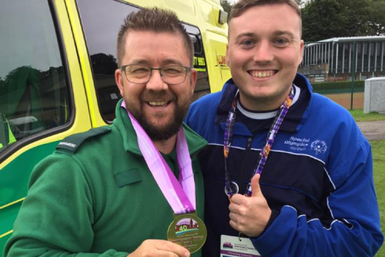 Celebrating Success at a Special Event: Two smiling men standing in front of an ambulance. One is wearing a green uniform and proudly displaying a medal, while the other is giving a thumbs-up, wearing a lanyard and event badge. The image captures a moment of joy and achievement at a community event.