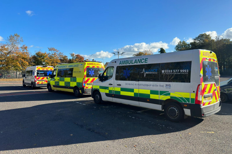 A row of Alliance Ambulance Services vehicles, marked with emergency and NHS partnership branding, are parked in a car park on a sunny day. The ambulances are yellow and white with green reflective markings, and trees with autumn leaves are visible in the background.