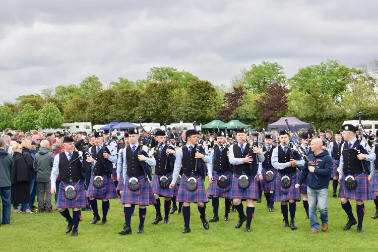 A group of musicians in traditional Scottish attire, including kilts and sporrans, perform in a bagpipe band at a Highland Games event, surrounded by a crowd of spectators under a cloudy sky.