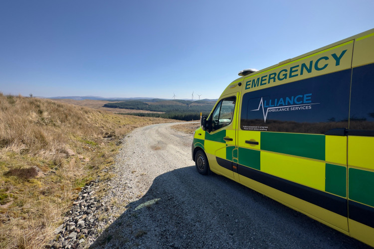 An Alliance Ambulance Services vehicle parked on a gravel track in a remote rural area, with open moorland, distant forests, and wind turbines on the horizon under a clear blue sky.