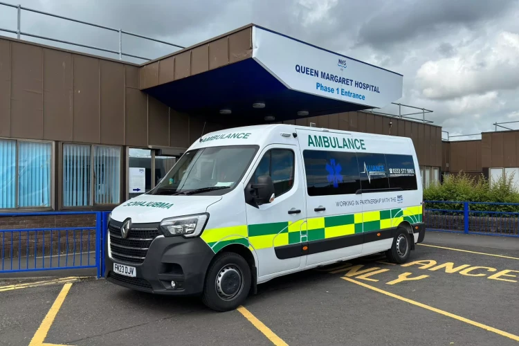 A white Alliance Ambulance Service vehicle is parked in the "Ambulance Only" zone outside the Phase 1 Entrance of Queen Margaret Hospital. The van features green and yellow checkered markings and the Alliance logo, indicating a partnership with the NHS. The hospital building, with its brown exterior and blue signage, is visible in the background under a cloudy sky.