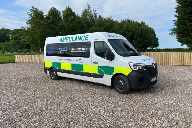 An Alliance Ambulance Service vehicle, marked with NHS Scotland branding and contact information, is parked on a gravel surface in an outdoor setting surrounded by trees and a wooden fence under a partly cloudy sky.